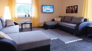 Una sala de estar con un sofá gris, una mesita, un televisor montado en la pared y una ventana con cortinas amarillas.