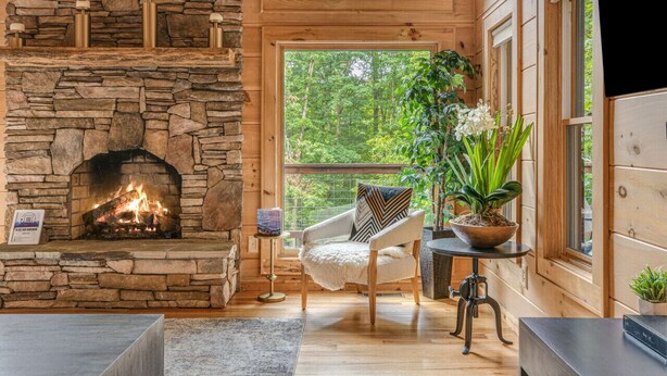 Una chimenea de piedra con fuego, una silla de madera y una planta en maceta en una habitación con ventanas amplias y paredes de madera.