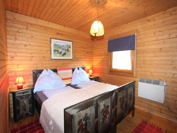 A wooden cabin bedroom with a bed, two bedside lamps, a window with a blind, a radiator, and a painting on the wall.