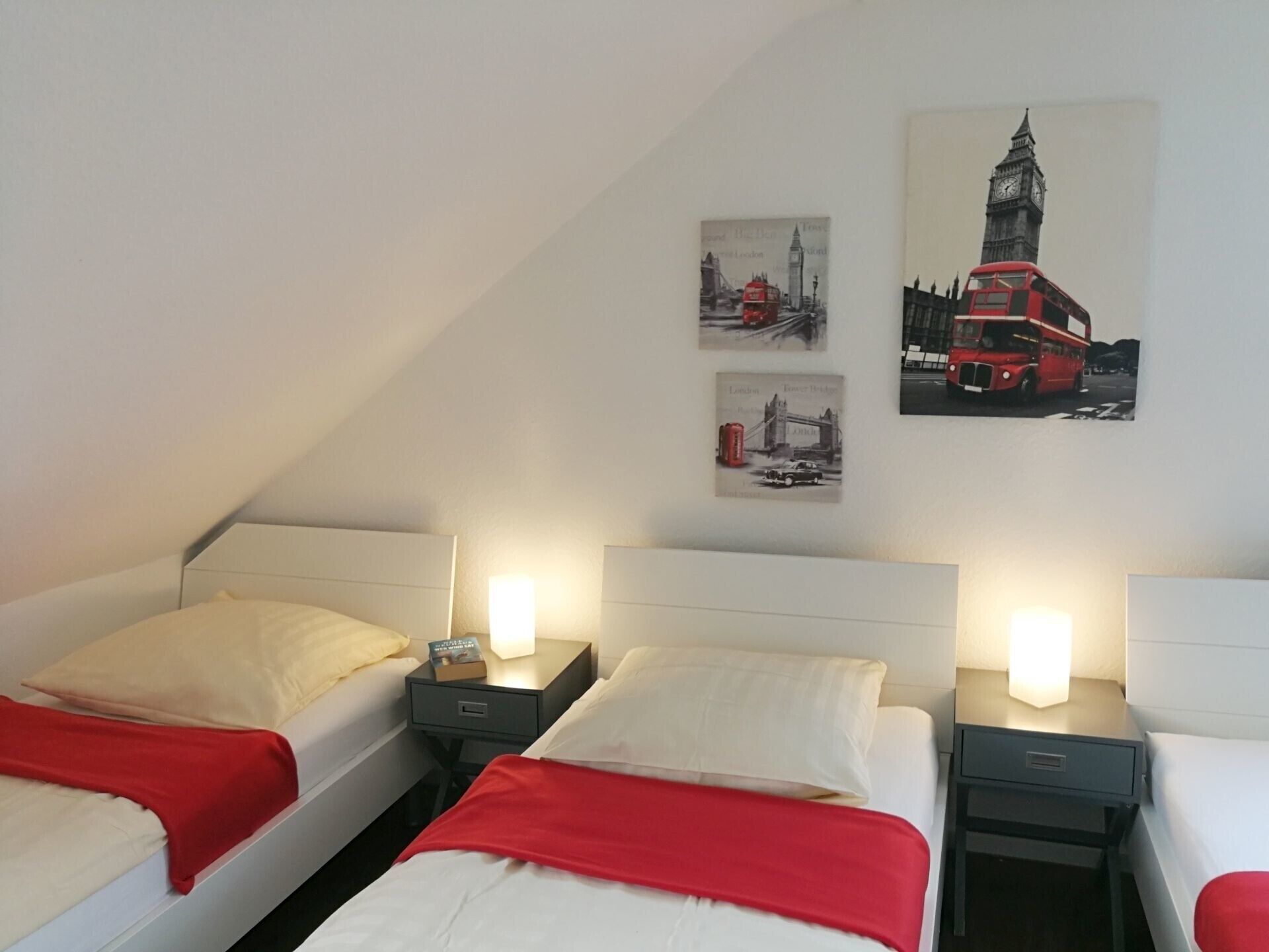 Ein Hotelzimmer mit zwei Betten, jeweils mit roter Decke, weißen Kissen und Nachttischen mit Lampen. An der Wand hängen drei gerahmte Bilder, die Londoner Sehenswürdigkeiten zeigen.