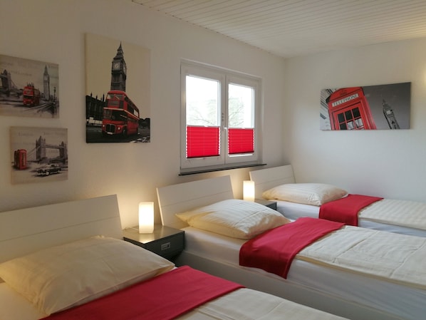 Une chambre d’hôtel avec trois lits, une fenêtre avec des stores rouges et des œuvres d’art murales représentant des monuments de Londres.