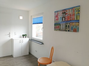Une petite pièce avec une fenêtre, une armoire blanche, une chaise et une œuvre d’art colorée accrochée au mur.