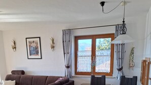 Een woonkamer met een bruine bank, een eettafel met stoelen, een raam met gordijnen en een aan de muur hangend schilderij.