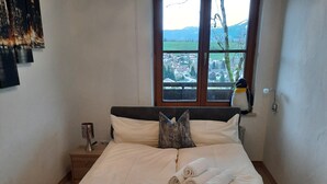 Een net opgemaakt bed met opgerolde handdoeken en een knuffel, een bedlampje en een raam met uitzicht op een stad en bergen.