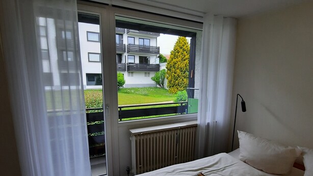 A bedroom with a bed, a radiator, a window with curtains, and a view of a building and greenery outside.