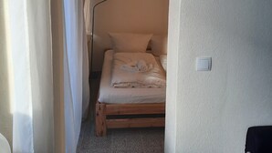 A narrow hallway leading to a bedroom with a wooden bed, white curtains, and a light switch on the wall.