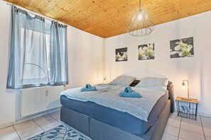 Un dormitorio con cama, un radiador, una ventana con cortinas, una lámpara de pared y tres cuadros de flores.