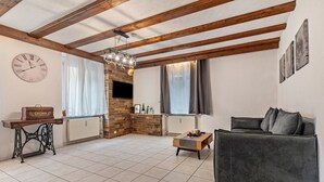 Una sala de estar con vigas de madera a la vista, una chimenea de ladrillo, un sofá, una mesa de centro, una máquina de coser antigua y un reloj en la pared.