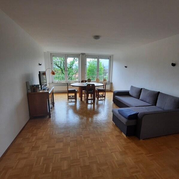 Un vaste espace de vie doté d’un parquet en bois, d’un canapé gris, d’une table à manger avec des chaises et de grandes fenêtres offrant une vue sur les arbres et les bâtiments.
