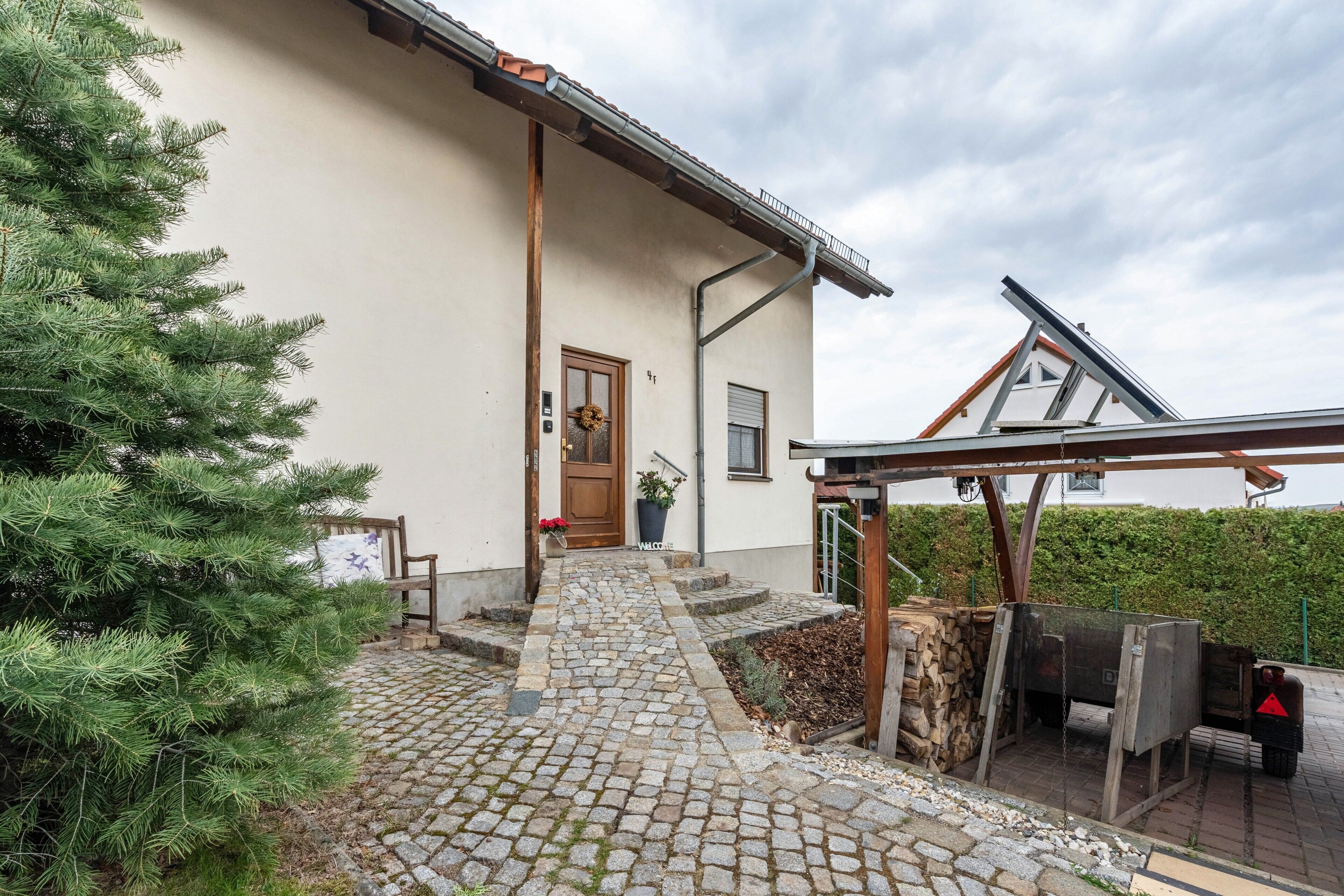 Ein Gebäude mit einem steinernen Weg, der zu einer Holztür führt, einem kleinen Garten mit einer Kiefer und einer Solaranlage.