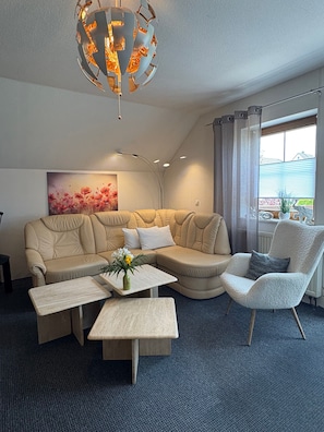 Un salon moderne avec un canapé d’angle, un fauteuil blanc, une table basse et un lustre original.