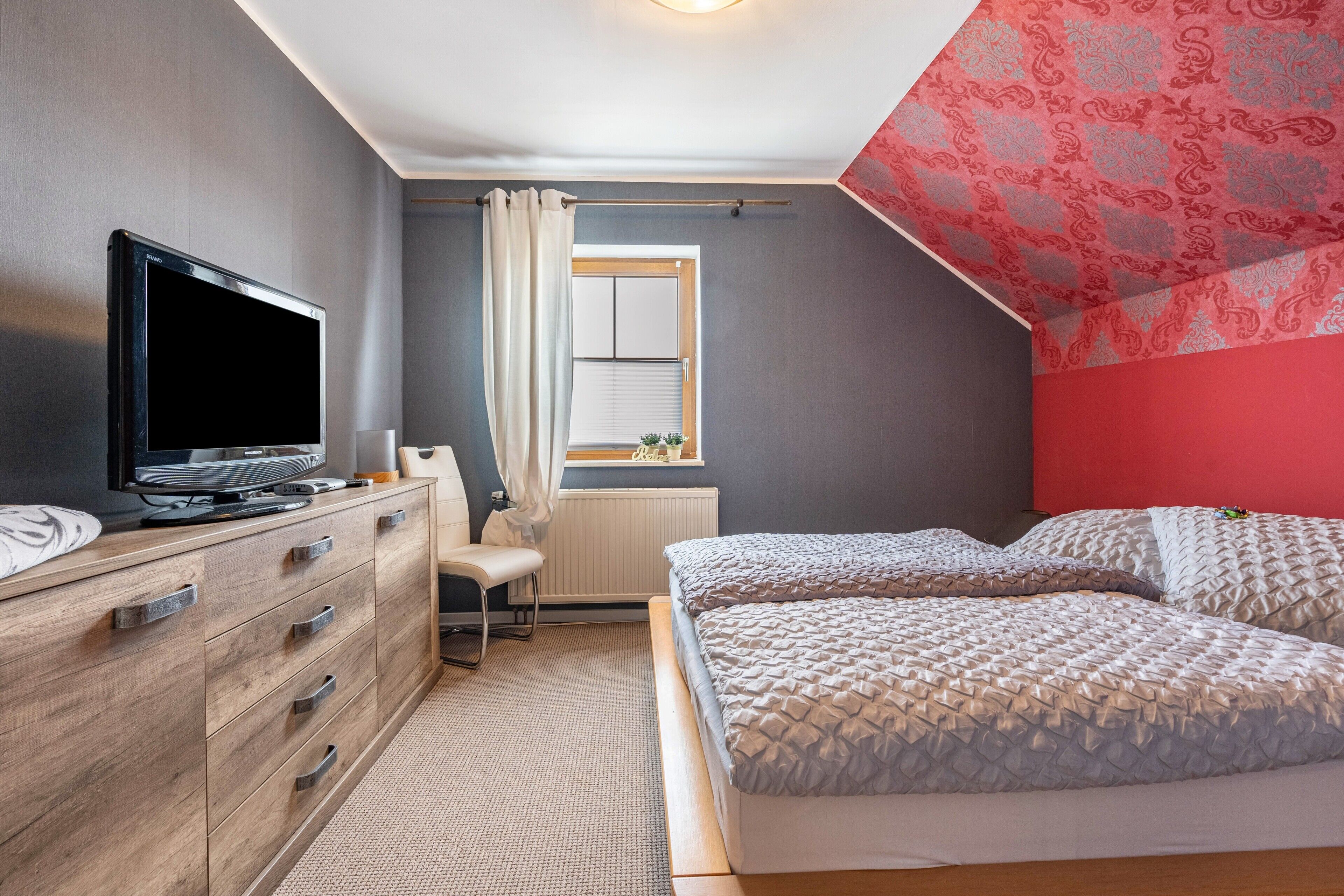 Un dormitorio con cama, televisión, cómoda y ventana con cortinas.