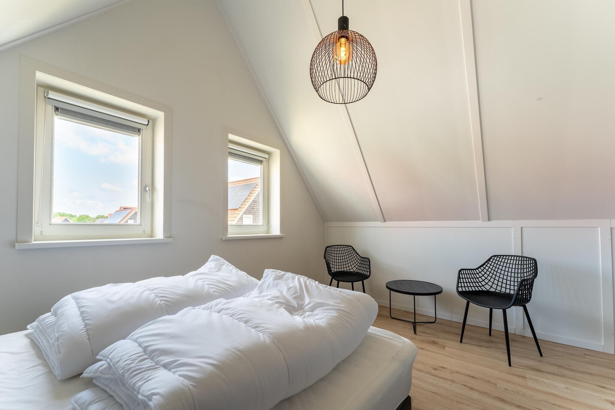 Et soveværelse med seng, to vinduer, en hængende lampe og to stole.