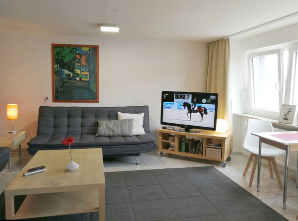 A living room with a grey sofa, a television displaying a horse riding event, a painting on the wall, and a window with curtains.