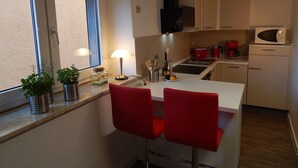 Een kleine keuken met een raam, een magnetron, een koffiezetapparaat en twee rode stoelen.
