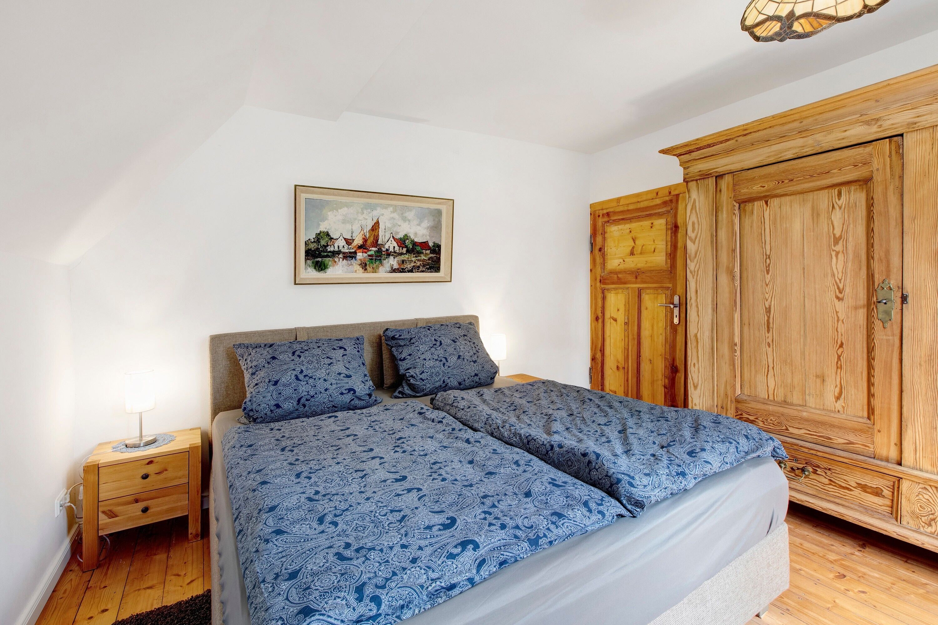 Un dormitorio con cama, mesita de noche, armario de madera y un cuadro en la pared.