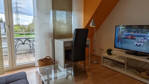 Una sala de estar moderna con una mesa de vidrio, una silla de oficina negra, un televisor mostrando una carrera y vistas al exterior a través de un ventanal.