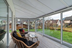 Une véranda meublée de mobilier en osier, dotée de grandes portes vitrées et offrant une vue sur un quartier résidentiel.