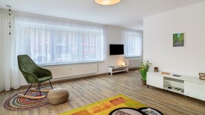 Un salon moderne avec un fauteuil vert, un téléviseur à écran plat, une console blanche et une grande fenêtre ornée de rideaux légers.