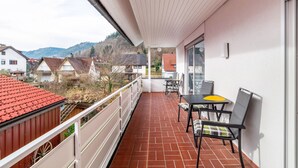 Un balcon doté d’un sol carrelé de tuiles rouges, une table avec des chaises, et une vue sur des maisons et des collines en arrière-plan.