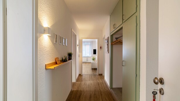 Un couloir avec un plancher en bois, un luminaire, une étagère avec des plantes et une porte dont la clé est dans la serrure.
