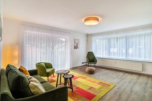 Uma sala de estar com um sofá verde, duas cadeiras, uma mesinha, um tapete colorido e janelas amplas com cortinas leves.