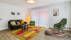 Un salon moderne avec un canapé vert, une petite table ronde, un fauteuil vert, un tapis coloré et une grande fenêtre ornée de rideaux légers.