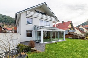 Une maison moderne à deux étages, dotée d’une véranda en verre, d’une pelouse bien entretenue et offrant une vue sur les montagnes.