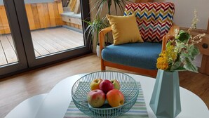 Een kamer met een houten vloer, een bank met een blauw kussen, een kussen met een chevrond patroon, een metalen schaal vol fruit en een vaas met bloemen.