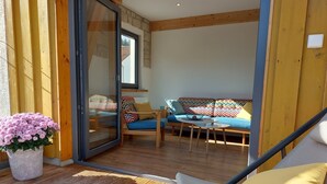 Een zonnig terras met een houten dek, een schuifpui naar een kamer met een bank en een tafel, en een potplant.