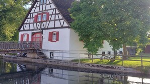 Een traditioneel vakwerkhuis met een houten brug over een gracht, omgeven door bomen.