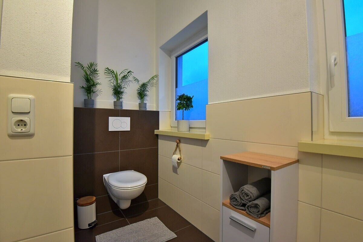 A small bathroom with a toilet, a window, a shelf with towels, and a trash bin.