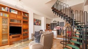 A living room with a sectional sofa, a TV, wooden bookshelves, and a modern staircase.
