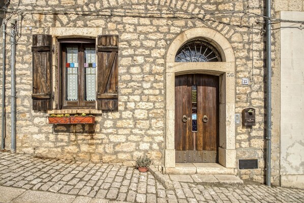 Un edificio de piedra con puerta y contraventanas de madera, suelo adoquinado y una pequeña planta en maceta.