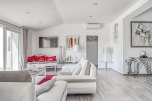Uma sala de estar moderna com um sofá branco, uma mesa de centro de vidro e uma almofada vermelha decorativa. Há uma TV montada na parede, um abajur e um quadro.