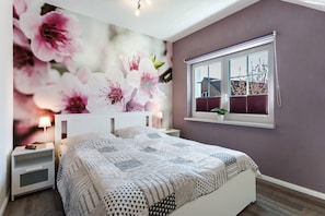 A bedroom with a bed, nightstands, a window with blinds, and a wall with a floral mural.