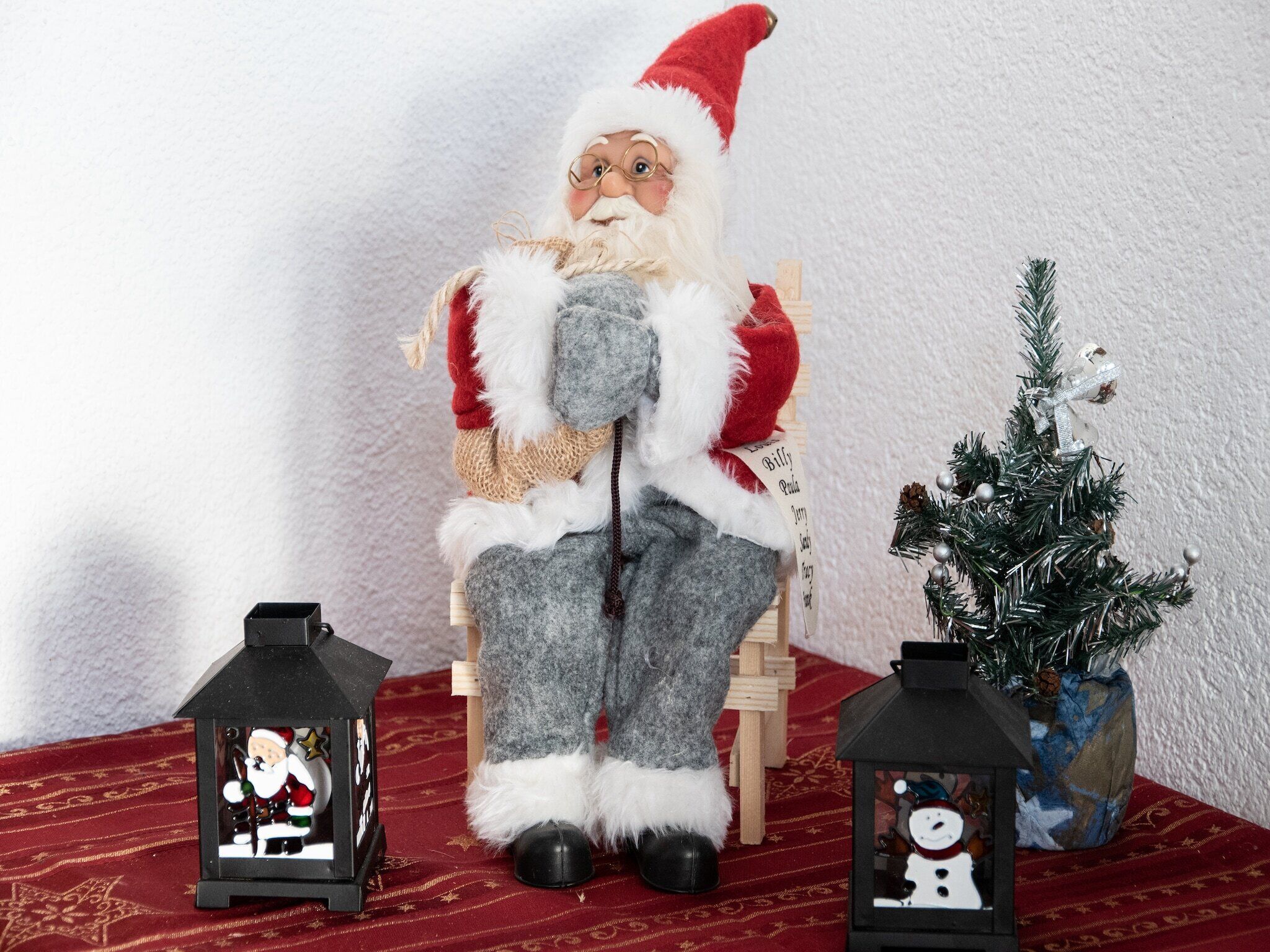 Eine Weihnachtselfe auf einem Stuhl, flankiert von zwei Laternen und einem kleinen Weihnachtsbaum.
