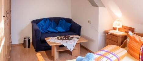 Una habitación acogedora con un sofá azul oscuro, una pequeña mesa de madera con bombones, una mesita de noche de madera con una lámpara y una cama colorida con toallas azules.