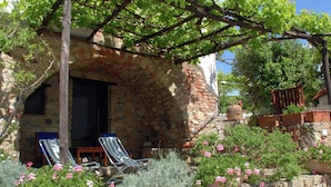 Ein Steingebäude mit einer Pergola, die in dichtes Grün rankt, zwei gestreifte Liegestühle und Kübelpflanzen mit rosa Blüten.