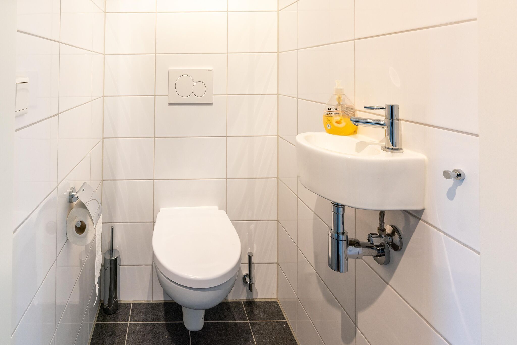 Ein kleines, modernes Badezimmer mit weißen Fliesen, einer weißen Toilette, einem wandmontierten Waschbecken mit Seifenspender und einem Toilettenpapierhalter.