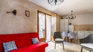 A living room with a red sofa, a white coffee table, and wicker chairs. There is a kitchen area with cabinets and a dining table.
