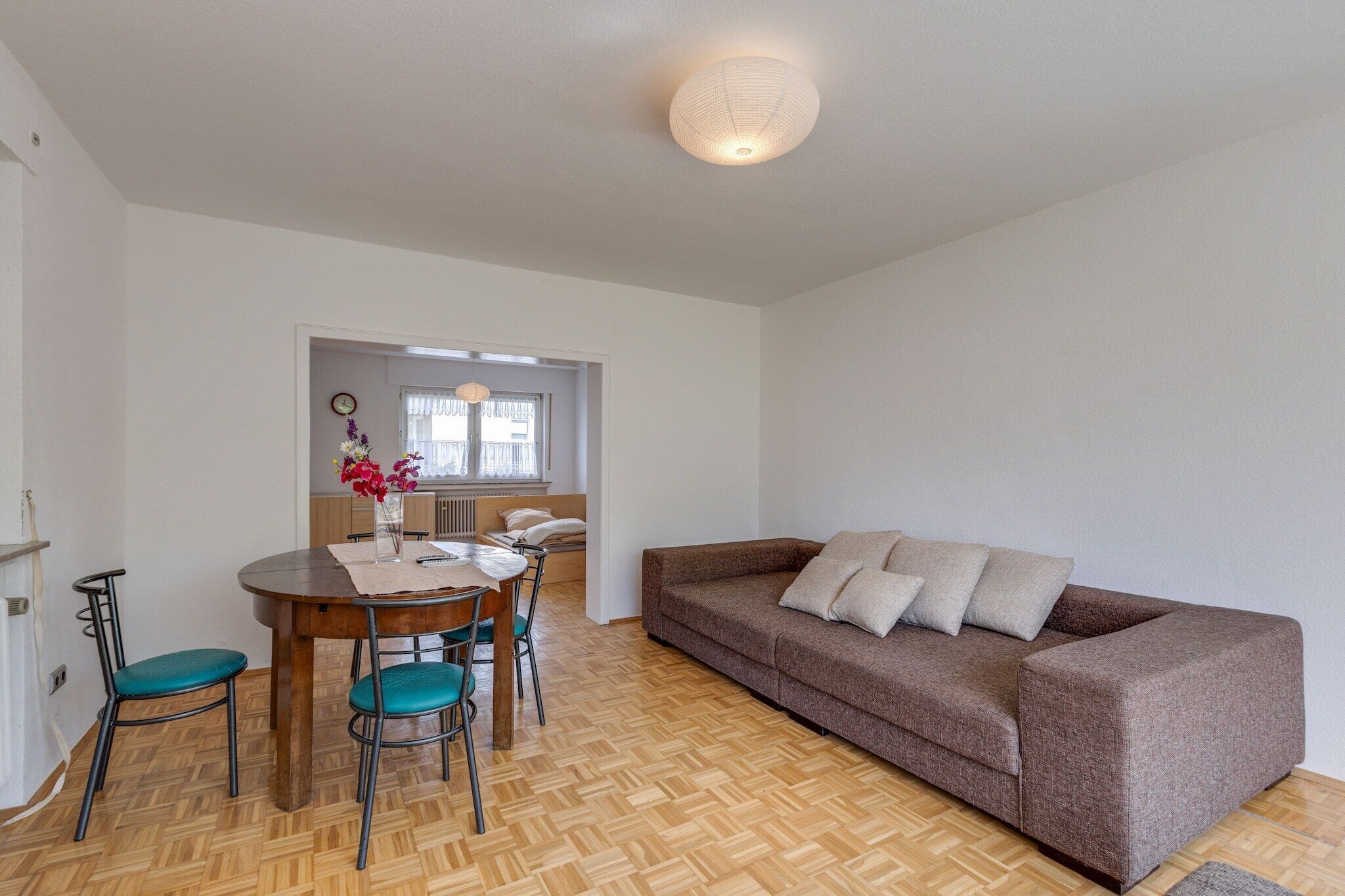 A room with a wooden herringbone floor, a brown sofa with cushions, a dining table with chairs, and a vase with flowers.