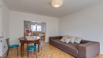 A room with a wooden herringbone floor, a brown sofa with cushions, a dining table with chairs, and a vase with flowers.