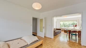 A modern room with a wooden floor, a bed with beige bedding, a dining table with chairs, and a window with a view of greenery.