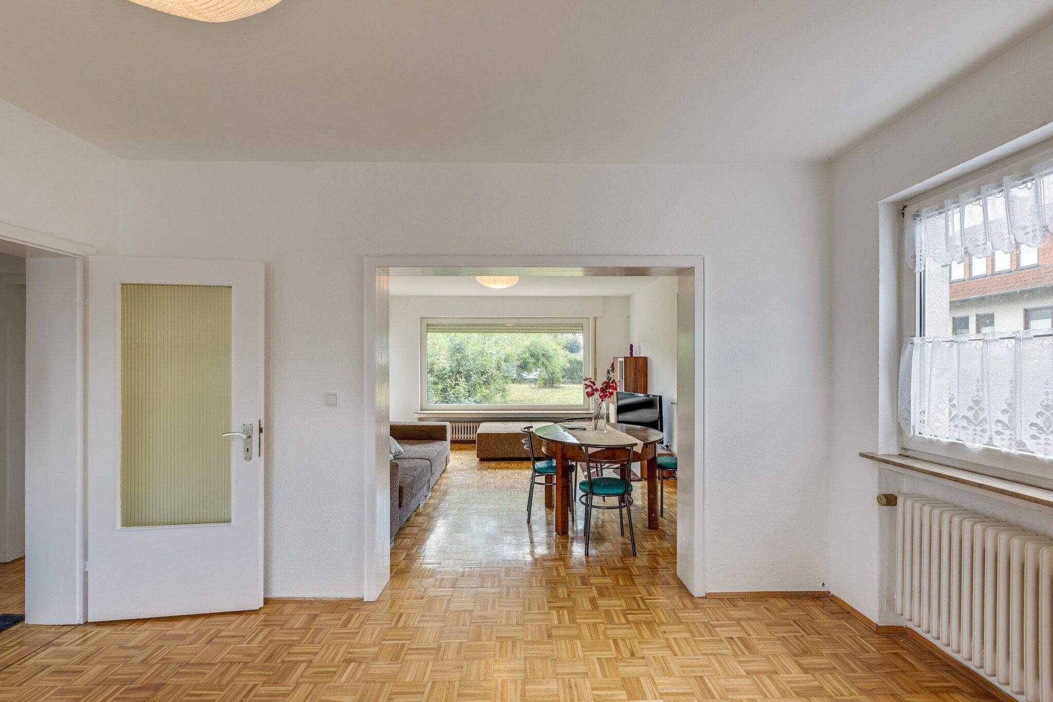 A spacious room with a wooden herringbone floor, a dining area with a table and chairs, a sofa, and a large window with a view of greenery.