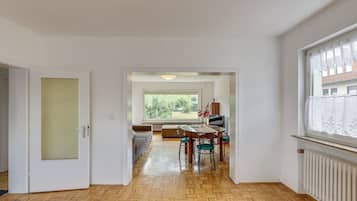 A spacious room with a wooden herringbone floor, a dining area with a table and chairs, a sofa, and a large window with a view of greenery.