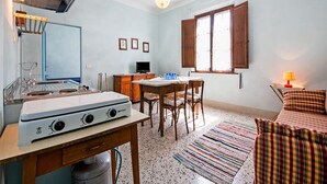 A compact kitchen with a gas stove, a sink, and a small dining area with a table and chairs. There is a window with wooden shutters, a ceiling light, and a television on a wooden cabinet.