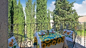 A terrace with a table set for two, surrounded by metal railings, overlooking a landscape of tall trees and a hill.
