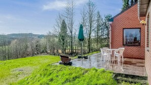 A patio with a table and chairs, a green umbrella, and a view of trees and hills.
