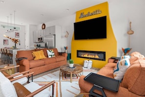 Un salon moderne avec une cheminée, un téléviseur fixé au mur et une cuisine en arrière-plan.